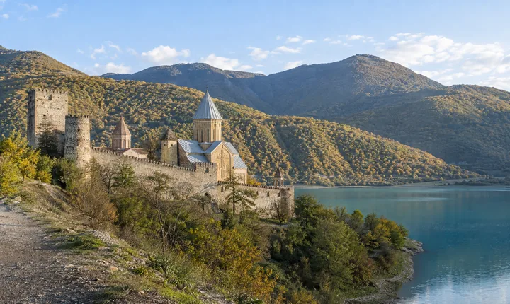 A stone fortress and domed church beside a turquoise reservoir and wooded hills