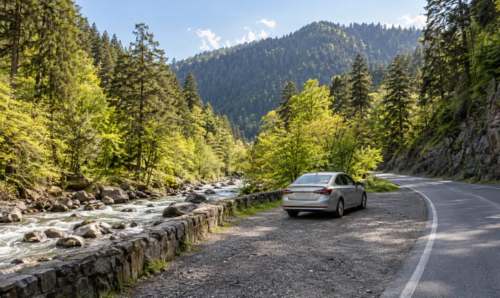 A silver sedan at a pull-off beside a paved road through a wooded Georgian river valley