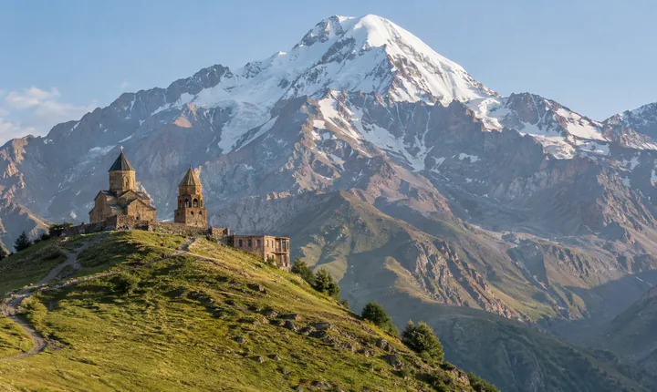 A stone church and bell tower stand on a grassy ridge below snow-covered Mount Kazbek