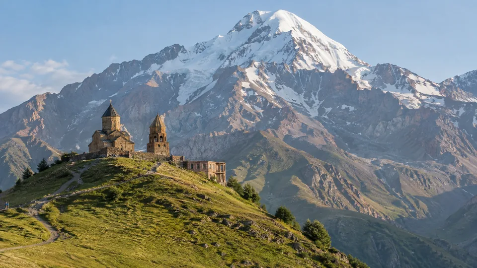 A stone church and bell tower stand on a grassy ridge below snow-covered Mount Kazbek