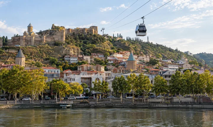 A cable car crosses above Tbilisi's river with hillside houses behind it