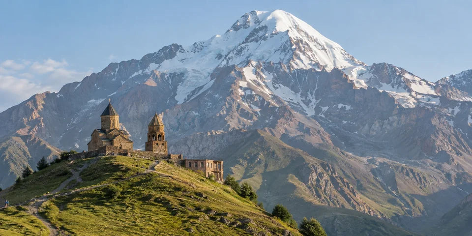 Eine Steinkirche und ein Glockenturm auf einem grasbewachsenen Bergrücken unter dem schneebedeckten Mount Kazbek